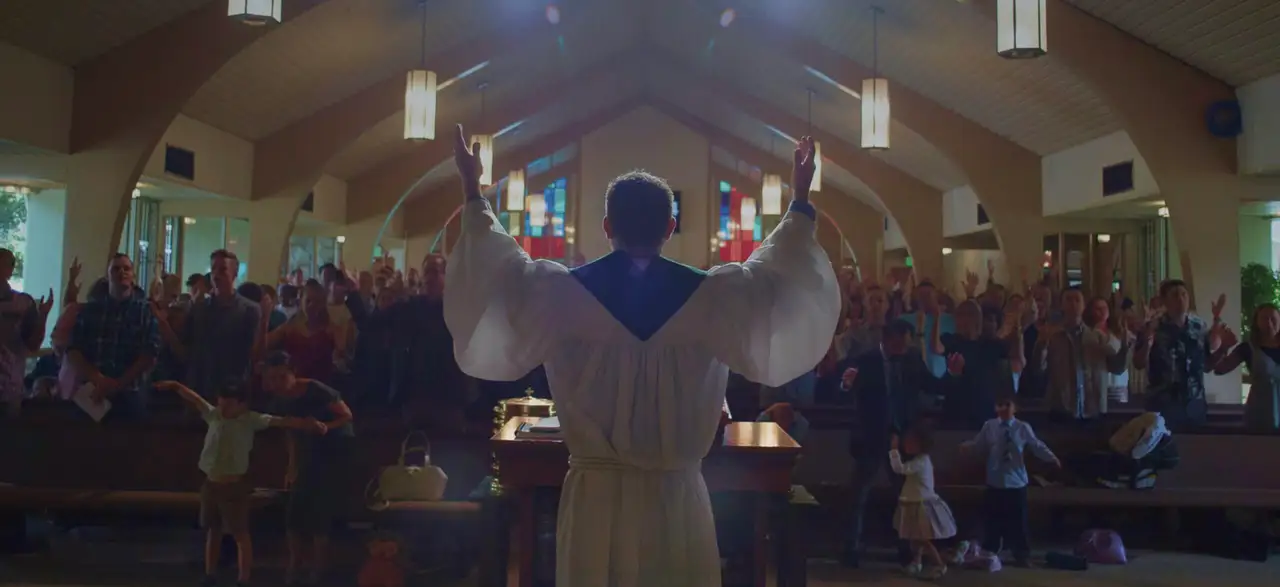 A minister in a white robe raises his hands in blessing over a congregation